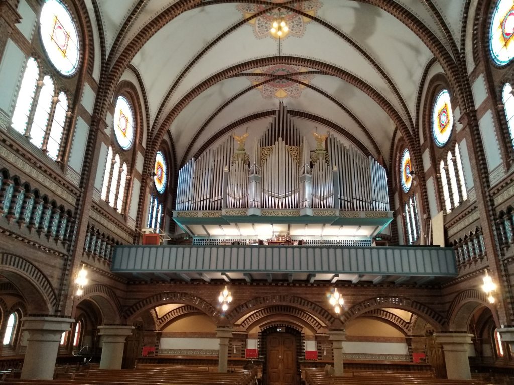 Blick aus dem Kirchenschiff auf die Orgelempore, die Orgel fült die Kirche fast in kompletter Breite, die Prospektpfeifen glänzen im Sonnenlicht